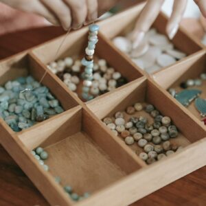 Crop unrecognizable female stringing beads on elastic cord while making bracelet at table with kit of assorted supply in room