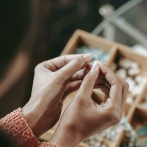 From above of crop faceless female stringing colorful bead on elastic rope while sitting at table with container of various supply