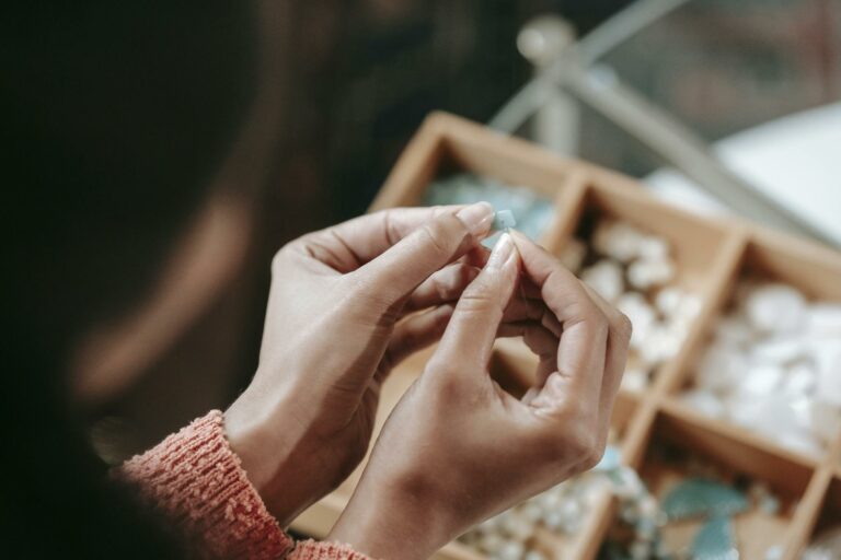 From above of crop faceless female stringing colorful bead on elastic rope while sitting at table with container of various supply