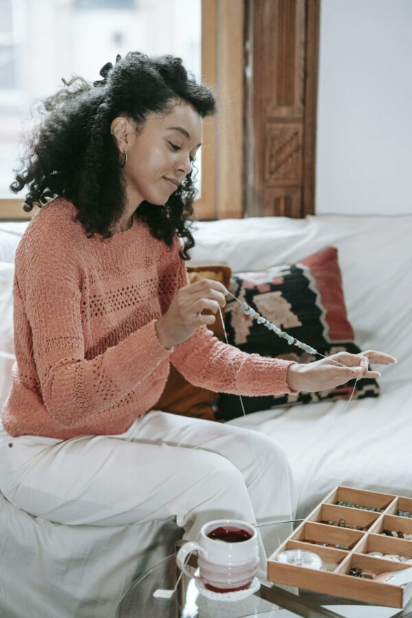 Side view of concentrated African American female with black hair stringing beads while making handmade bracelet while sitting in couch