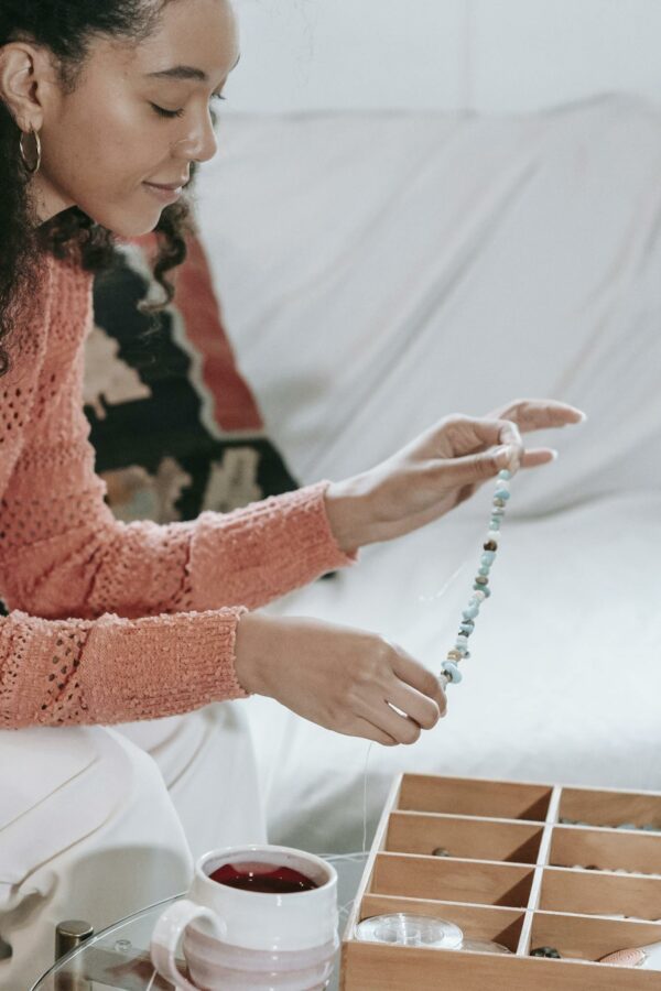 Side view of crop African American female with stringed beads in hands sitting at table with container of supply while making bracelet